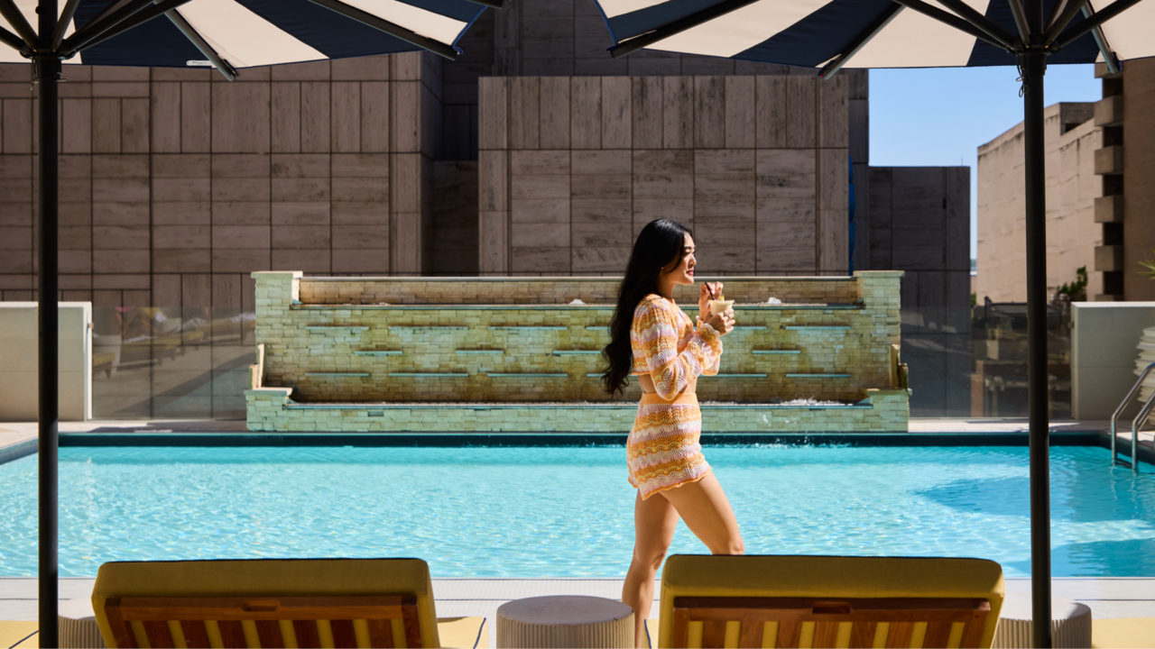 Woman sipping a drink walking by a pool with two umbrellas and pool chairs